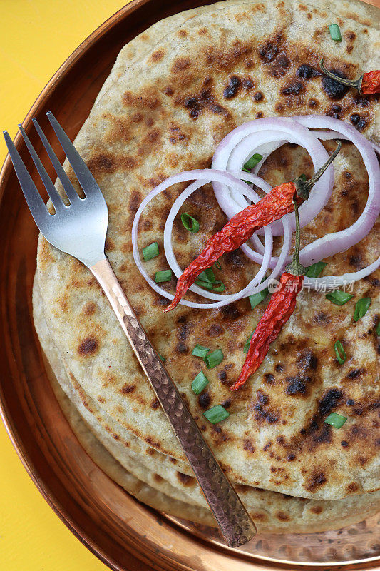 金属托盘上放着aloo paratha，上面撒着辣椒、红洋葱圈和切碎的韭菜，美味的面包里塞满了捣碎的garam masala土豆和金属叉子，黄色的背景，俯瞰的风景